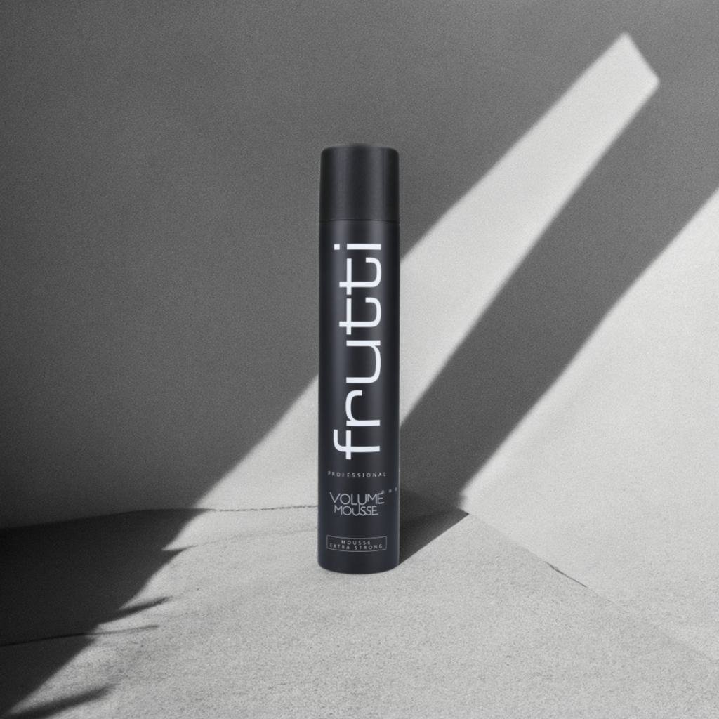 Czarna butelka pianki zwiększającej objętość Frutti Professional Volume Mousse, z białym logo i tekstem, umieszczona na betonowym podłożu z ciekawym grą światła i cienia tworzącym geometryczne wzory